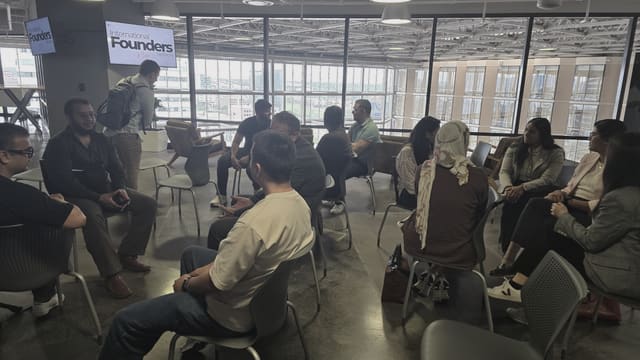 About a dozen people seated in a wide circle in conversation, with daylight coming through a full height glass wall behind them and an IFN meetup slide on a screen at the left.