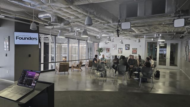 The meetup seen from behind a laptop on a stand at the front of the room, the group seated in a circle in the distance across a wide polished concrete floor.