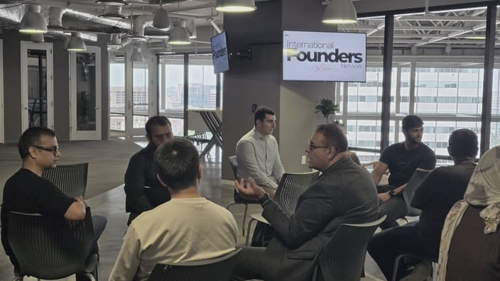 One person mid gesture speaking to a small seated circle, the others turned toward them, with an IFN meetup slide on the screen behind and the city through the glass.