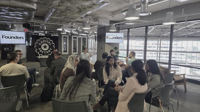 A seated circle of people listening, several of them in the foreground with their backs to the camera, with the room's windows and IFN screens behind.