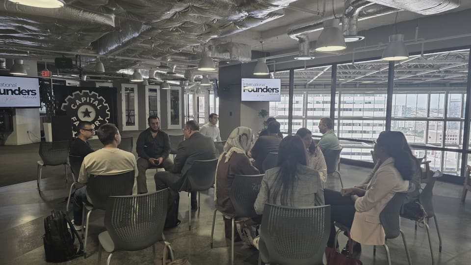 A circle of people talking across a wide open floor, photographed from behind the back row, with IFN meetup slides on two screens and downtown Austin through the windows.