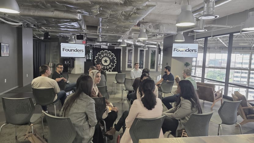 A full circle of about fifteen people seated in conversation on an open floor, several of them turned toward the camera, with IFN meetup slides on two screens and downtown Austin through a full height glass wall.