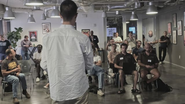 A speaker seen from behind addressing a mixed seated and standing crowd across a wide open floor under industrial pendant lights.