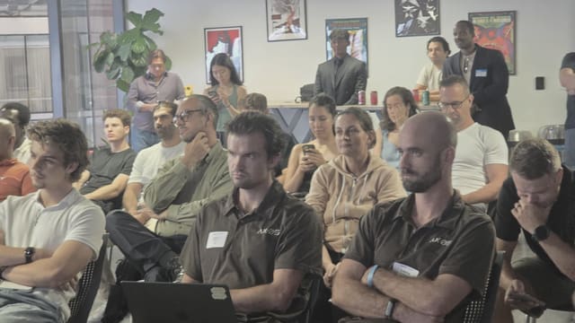 People seated in rows listening, some with laptops open on their laps, others standing along the back wall of a daylit room with framed posters.