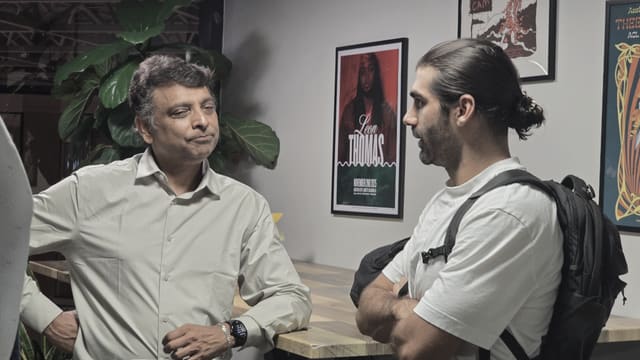 Two people talking across a counter in front of framed posters, with a leafy plant beside them.