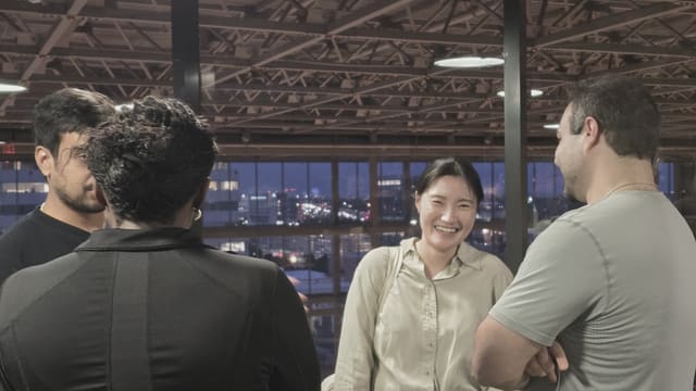 A small group talking and smiling beside tall windows with a city skyline at dusk behind them.