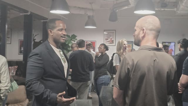 Two people mid conversation in a room of hanging pendant lights, one gesturing with both hands, with other groups talking behind them.