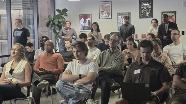 A seated audience turned toward a speaker out of frame, one person smiling in the foreground, with others standing along the back wall beside a tall plant.
