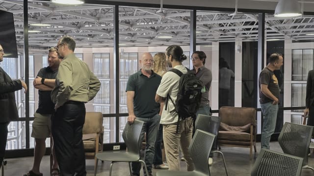 People standing in conversation near leather chairs and tall windows, with grey stackable chairs scattered across the polished floor.