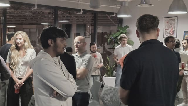 People standing and talking under industrial pendant lights, with a tall plant and glass partitions behind them.