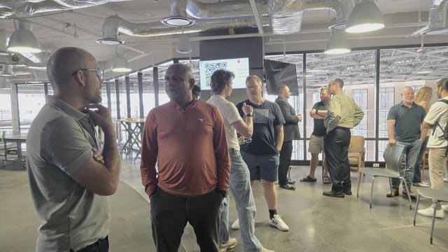 Small groups of people talking across an open floor, with a QR code lit on a pillar screen and the city through the windows behind them.