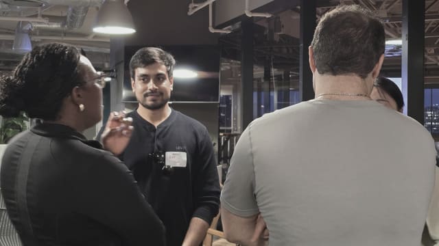A few people standing in conversation in a nightlit room, one wearing a name tag, with city lights visible through the windows behind them.