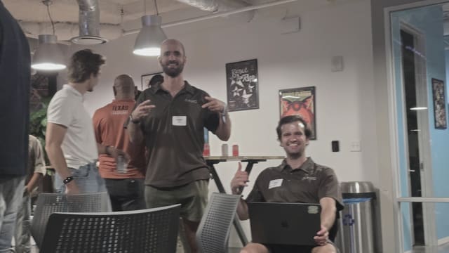 Two people facing the camera mid celebration in a meetup room, one seated with a laptop, others talking further back near framed posters.