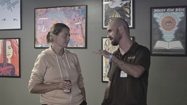 Two people standing and talking in front of a wall of framed posters, one of them holding a drink.