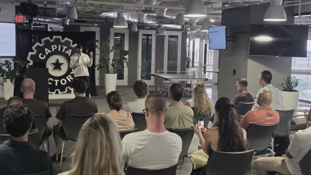 A speaker with a microphone facing a seated audience across an open floor, photographed from the back of the room, with a meetup slide on a screen and a wall emblem behind.