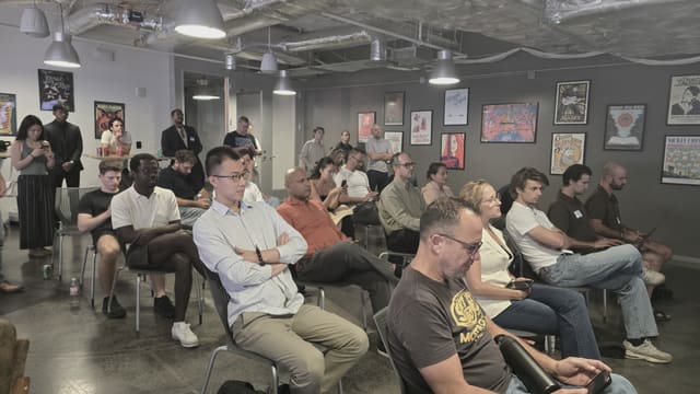 Rows of people seated and listening in an industrial room under hanging pendant lamps, with a few people standing along the back wall of framed posters.