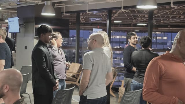 Small groups of people networking beside tall windows looking out onto a city skyline at dusk under hanging pendant lights.