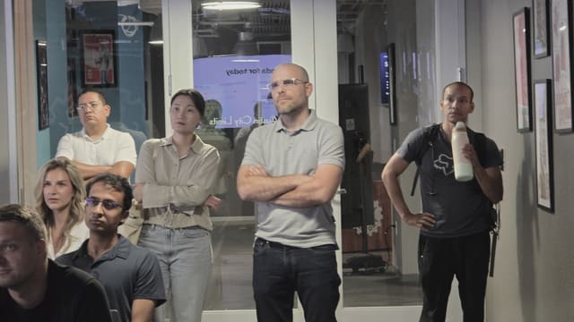 Several people standing and seated near glass doors listening, one holding a water bottle, with framed posters on the wall beside them.