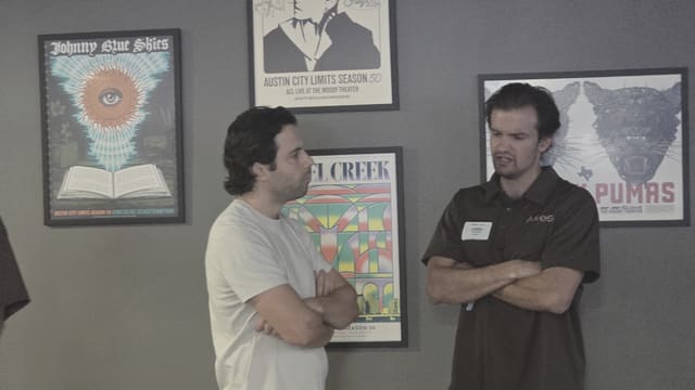 Two people standing with arms crossed in conversation in front of framed posters on a grey wall.