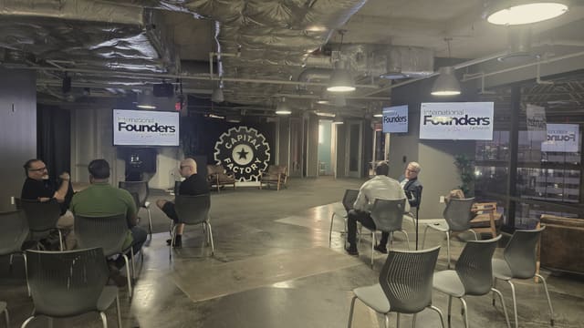 Two small groups of people talking across a large open room with rows of empty chairs around them, under a steel and ductwork ceiling, with lit IFN meetup slides on the screens and the city lights at the windows.