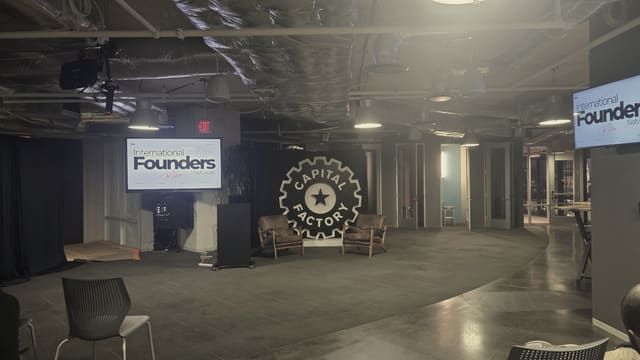 Two leather armchairs set in front of a large illuminated wall emblem at night, an IFN meetup slide on a screen to each side, empty chairs across the polished floor and one person seated at the edge of the frame.