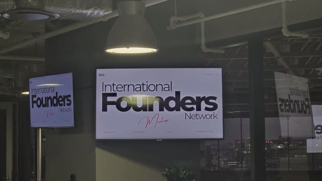 An IFN meetup slide lit on a wall mounted screen at night under a pendant lamp, with a second screen to the left and the slide reflected twice in the window glass over the city.