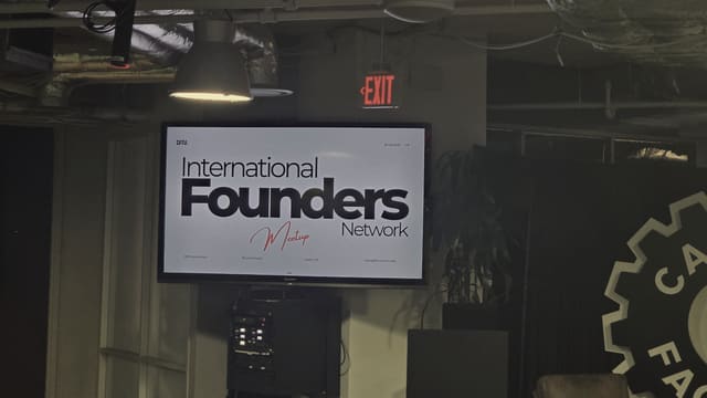 An IFN meetup slide lit on a wall mounted screen at night, a pendant lamp above it, a red exit sign to the right and the edge of a large wall emblem at the frame's edge.