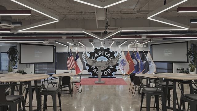 An empty event hall of high tables and stools before the evening starts, with national and military service flags standing either side of a large illuminated wall emblem and a projection screen at each end of the room.