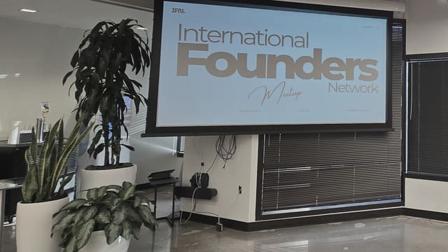 An IFN meetup slide projected on a pull down screen in a quiet corner of the room, two potted plants beside it and the ceiling lights reflected in the polished floor.