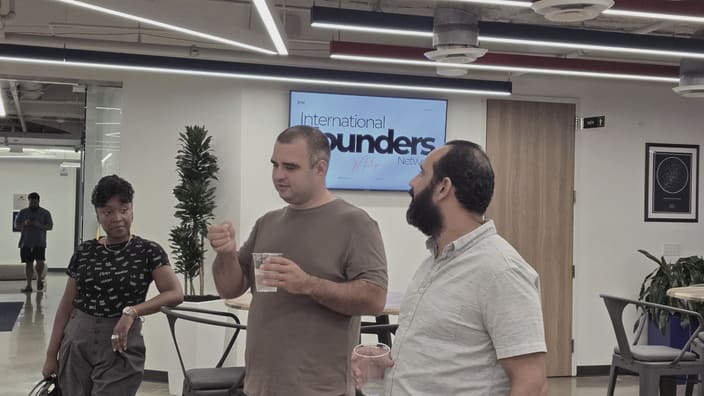 Three people standing and talking in an open room, two of them holding drinks, an IFN meetup slide on the screen behind them and another person standing further back beside the wall.