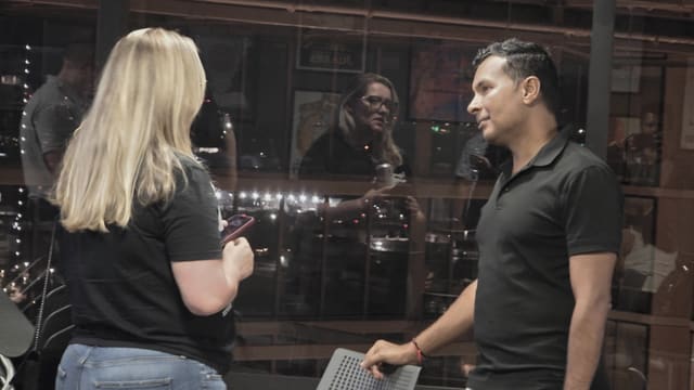 A man in a black polo and a woman with long blonde hair talking beside a glass window reflecting the room at night.