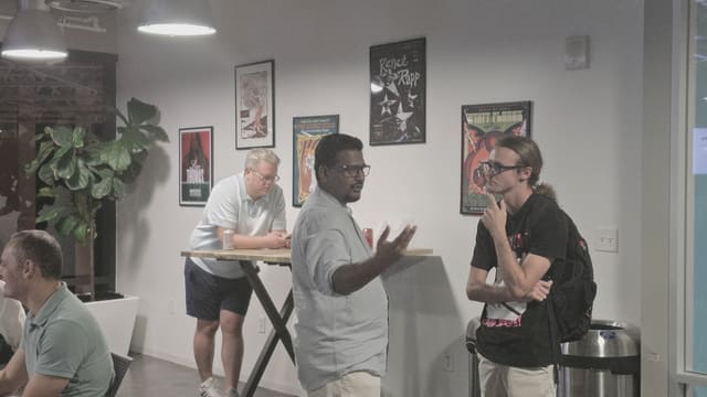 A man in a light blue shirt gesturing while talking with a young attendee wearing a backpack, posters and a plant behind them.