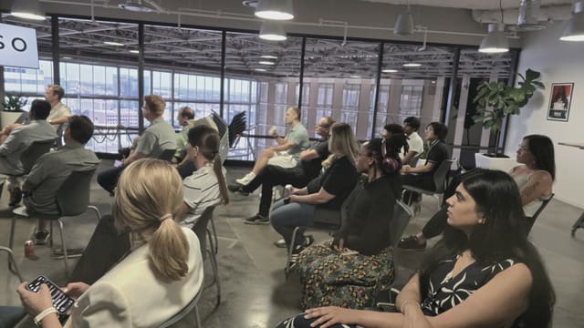Attendees seated facing a presentation, with floor-to-ceiling windows showing a city skyline at dusk behind them.