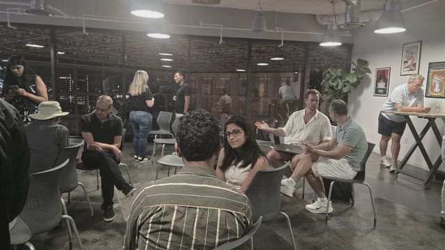 People networking across a nightlit room: some seated mid-conversation, others standing by tall windows overlooking city lights.