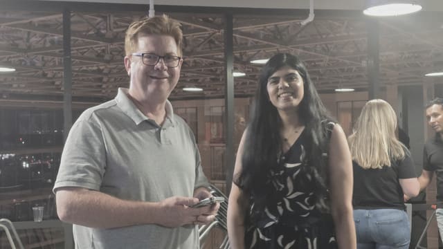 A man in a gray polo holding a phone and a woman in a patterned top smiling for the camera near stacked chairs and night windows.