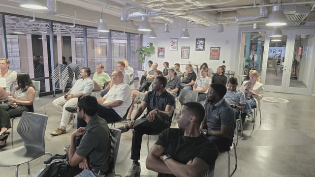Rows of attendees seated in gray chairs listening in a bright industrial meetup room with exposed ducts and framed posters on the wall.