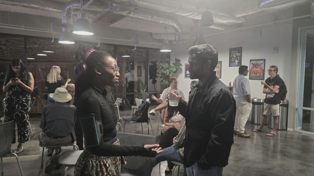 A woman with purple headphones gesturing as she talks with a man holding a drink, other attendees mingling behind them.